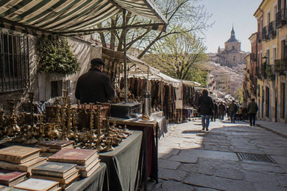 Flea market stalls with vintage goods and antiques displayed on tables along a sloped Madrid street