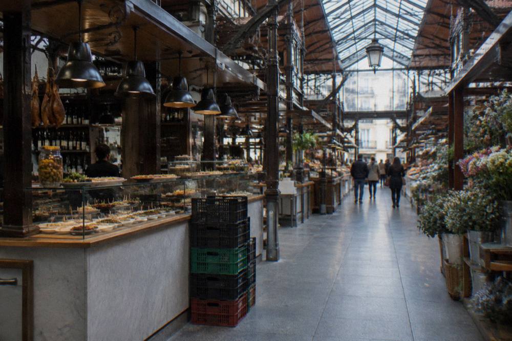 Interior of a glass-roofed market hall with food stalls and hanging lights along the aisles