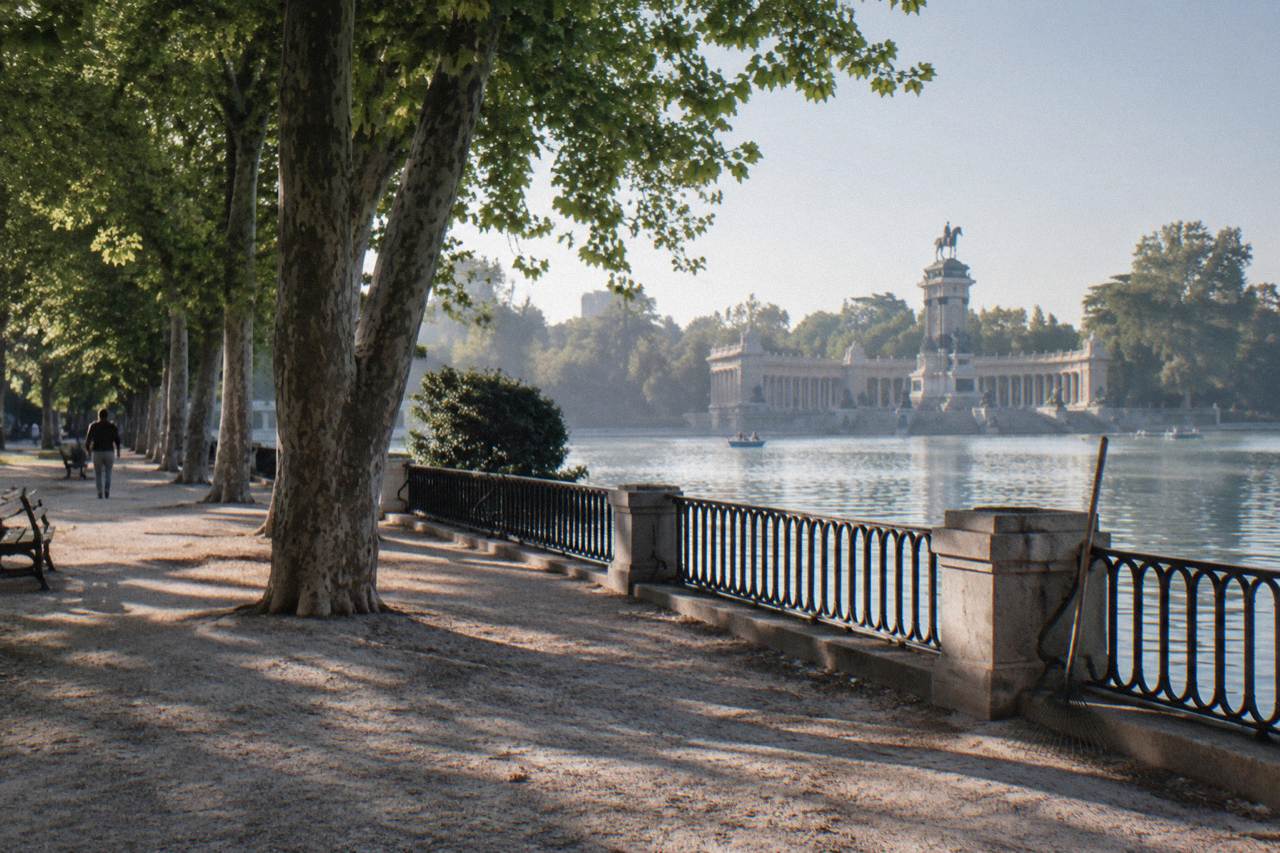 Tree-lined gravel path beside the lake in Retiro Park with the monument to Alfonso XII in the distance