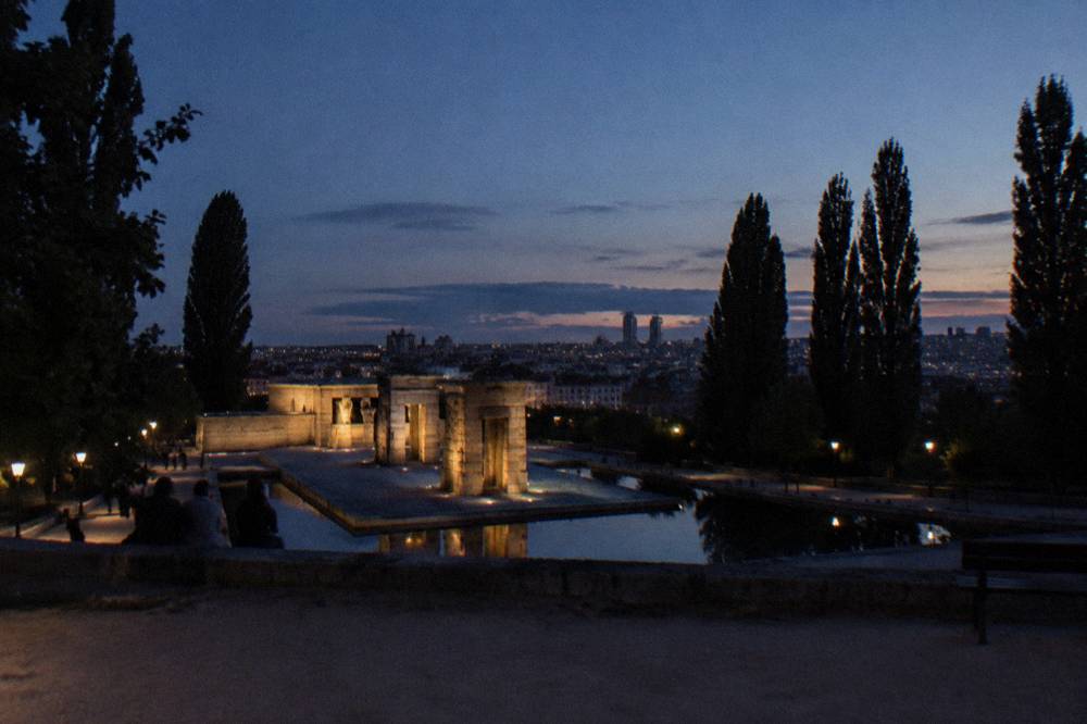 Wide view of a park at dusk with silhouetted trees and a city skyline glowing beyond a hilltop temple